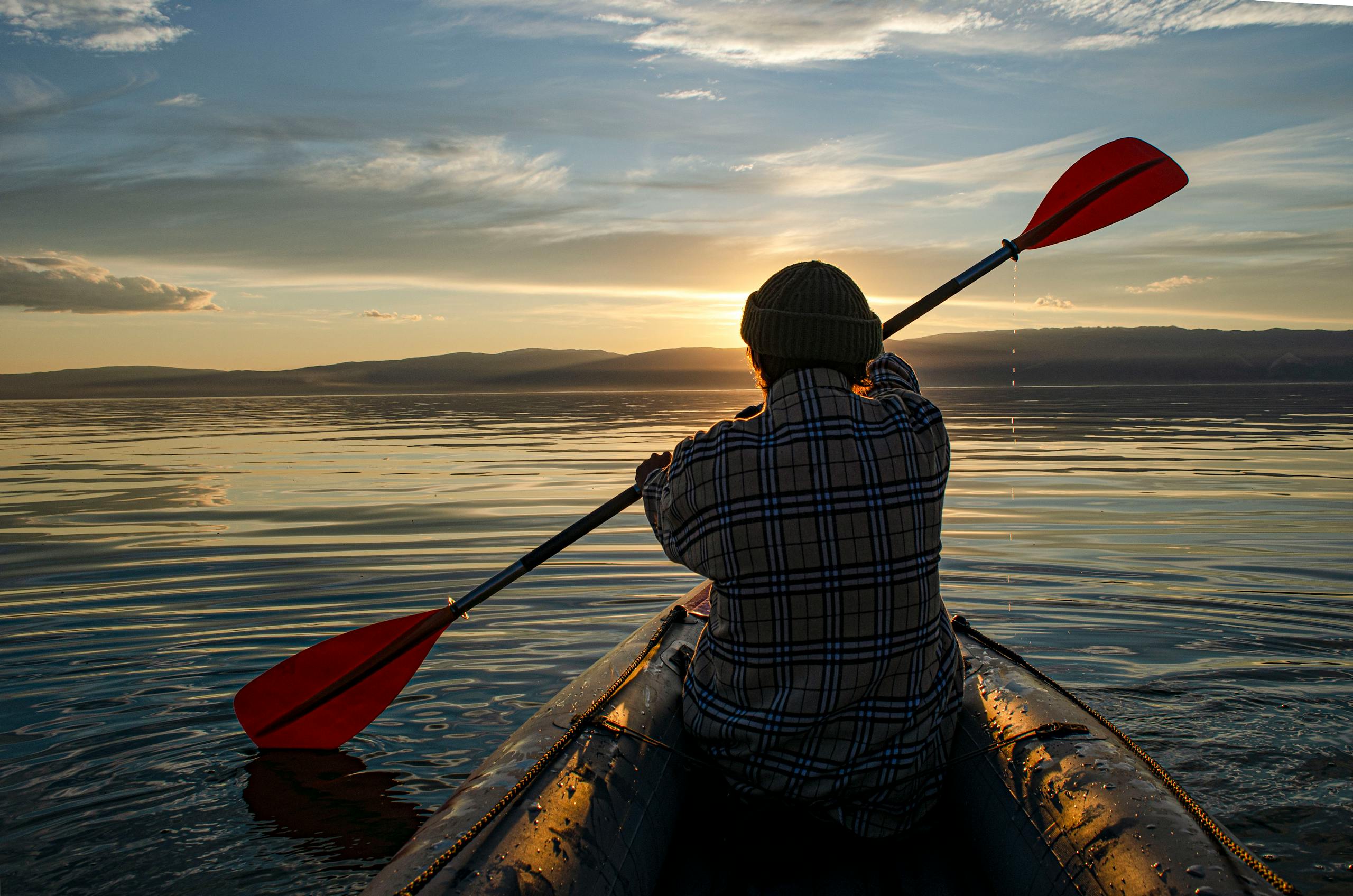 Person kayaking at sunset with plaid shirt and beanie on calm water